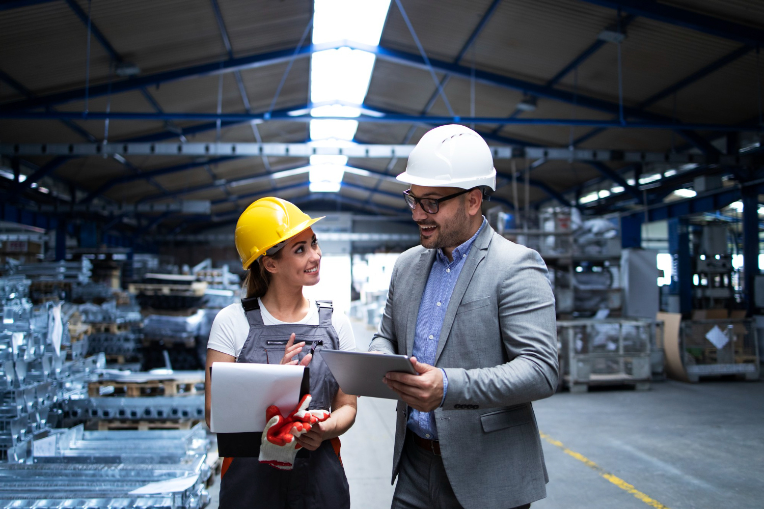 A Woman In A Yellow Hard Hat And Work Overalls Holding Documents And A Man In A Suit Holding A Tablet, Having A Discussion Inside A Large Industrial Warehouse