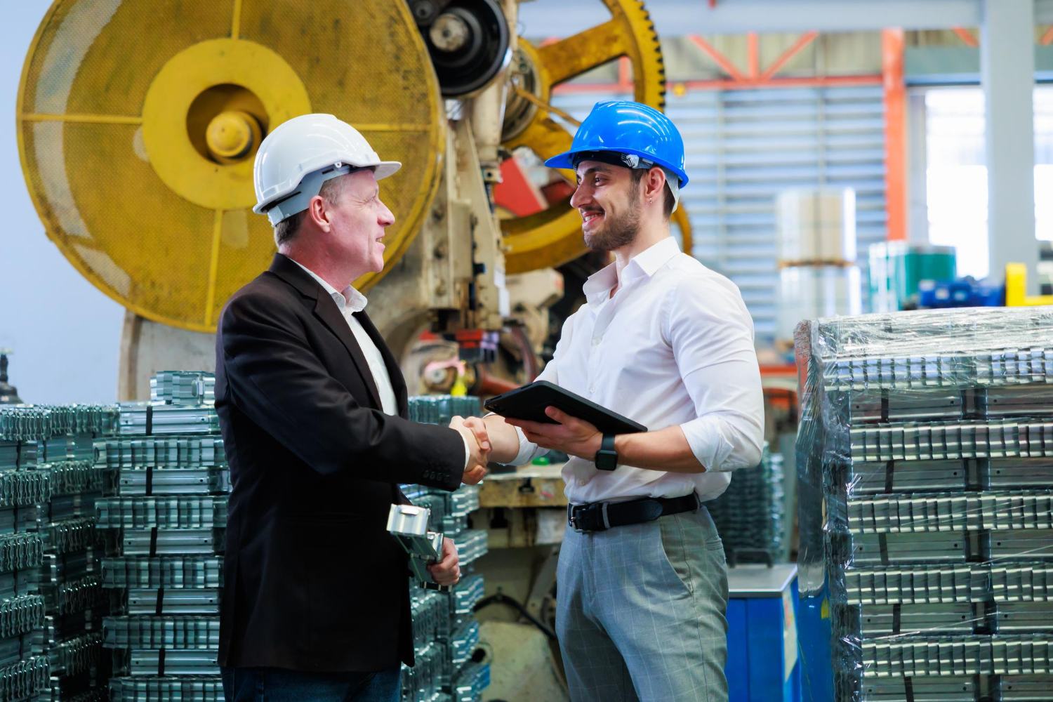 Two Men Wearing Safety Helmets Shaking Hands In An Industrial Warehouse Filled With Stacked Metal Materials, Symbolizing Agreement Or Partnership