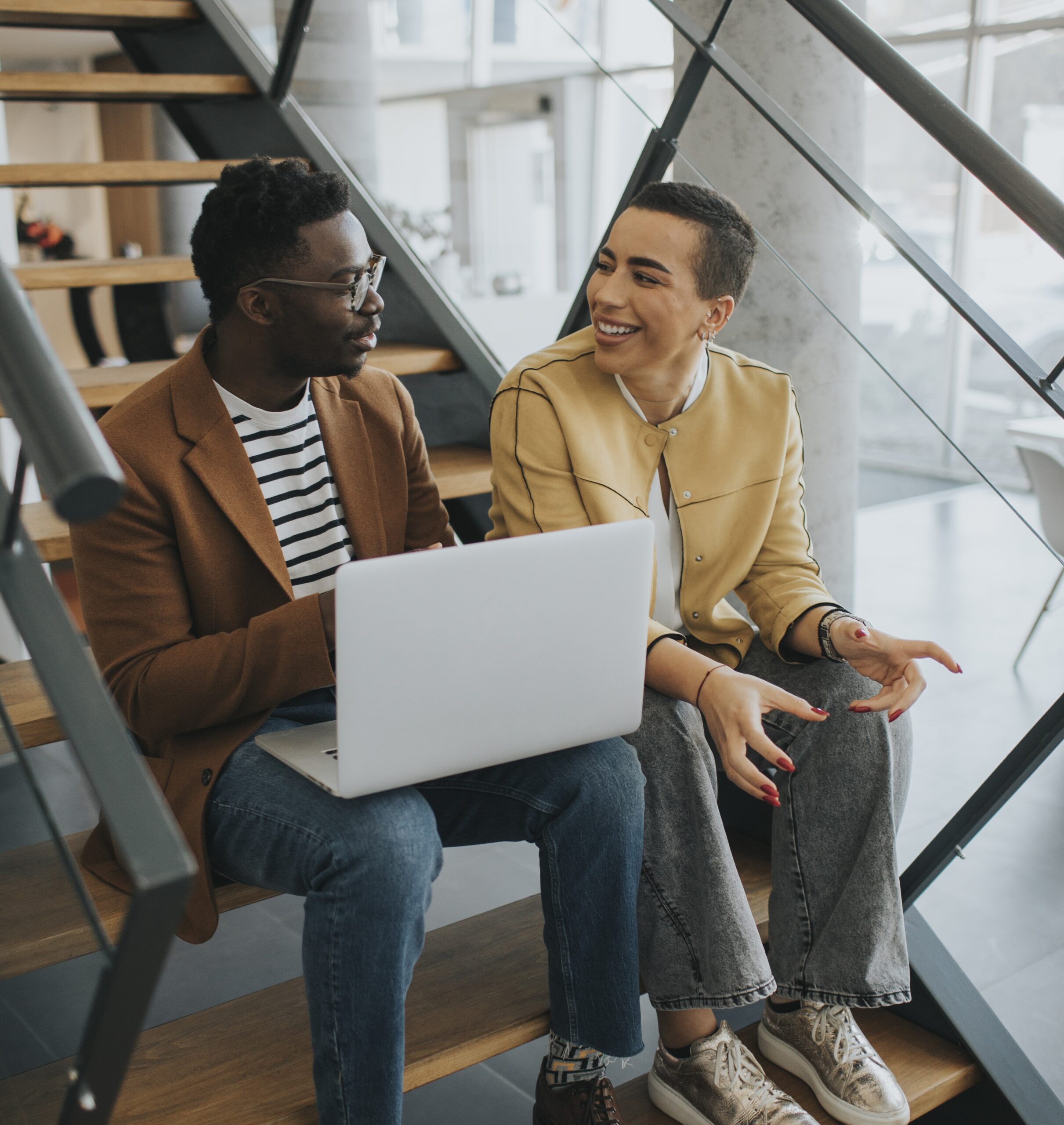 Two Young Professionals Sitting On Office Stairs, Engaged In A Friendly Conversation While Using A Laptop, With Natural Light Coming Through Large Windows